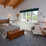 Bedroom with white walls, carpeting, wooden picket fence headboard, multicolored bedding, wooden trunk, and sitting area