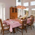 Dining room with white walls, carpeting, wooden table with pink tablecloth, wooden chairs, wooden china hutch, and many windows with views of the outside