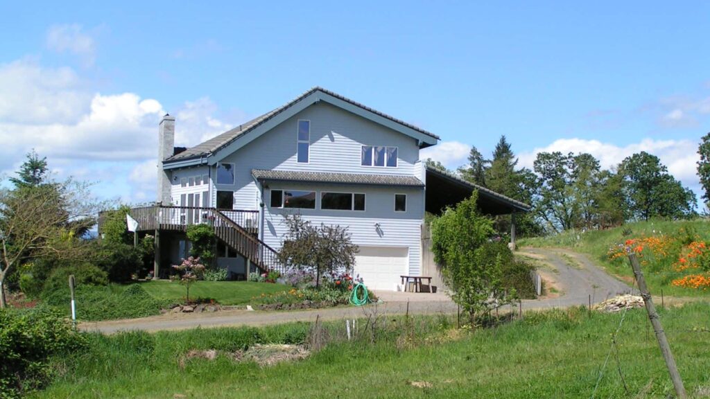 Exterior view of property painted light blue with white trim surrounded by green grass, trees, shrubs, and flowers